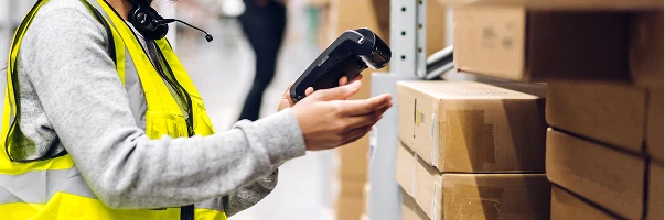 A worker in a yellow safety vest scans packages on a warehouse shelf with a handheld barcode scanner at Rexel Industrial.