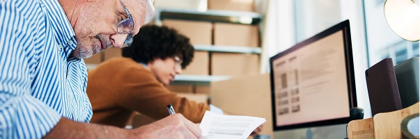 An older man in glasses writes on a document at a desk, whilst a younger person works at a computer in the background. Shelves with cardboard boxes are visible behind them, suggesting a Rexel Industrial office environment.