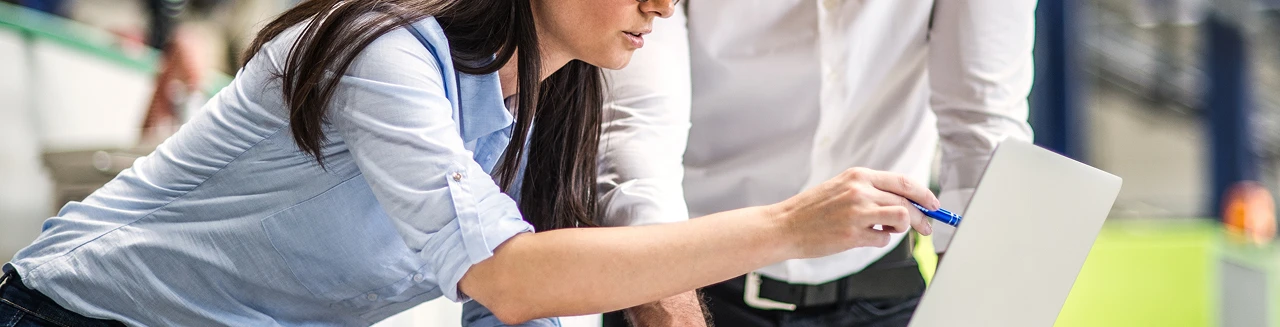 Two people stand close together, looking at a laptop screen. One person points at the screen with a pen, suggesting discussion or collaboration in a Rexel Industrial work setting.
